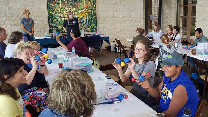 Photo of a group of about 25 teachers around a U shaped group of tables holding CO2 molecules made from pipe cleaners and styrofoam balls