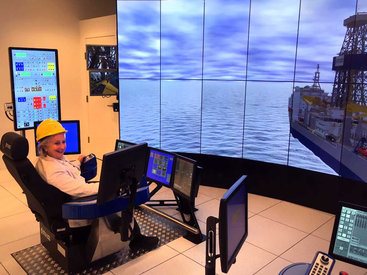Sue sits in front of monitors and a large screen (simulator) that shows a drilling rig out on sea